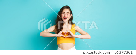 Summer holidays and emotions concept. Young romantic girl showing I love you heart gesture and smiling, standing over blue background 95377361