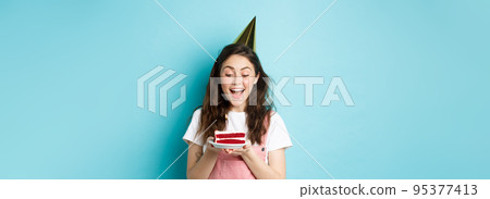 Holidays and celebration. Excited woman celebrating birthday, blowing candle on cake, wearing party cake and having fun, standing over blue background Holidays and celebration. Excited woman celebrating birthday, blowing candle on cake, wearing party cake and having fun, standing over blue background 95377413