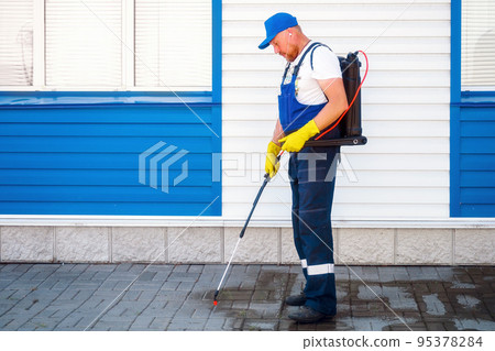 Man in work jumpsuit treats sidewalk from weeds. Disinfection of adjacent territory of house Man in work jumpsuit treats sidewalk from weeds. Disinfection of adjacent territory of house 95378284