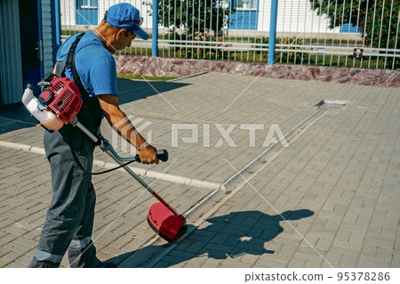 Janitor trims grass with trimmer on summer day on territory of production base. Man in work jumpsuit Janitor trims grass with trimmer on summer day on territory of production base. Man in work jumpsuit 95378286