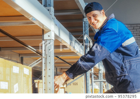 Storekeeper puts box on rack shelf looks at camera and smiles. Young Caucasian warehouse worker in Storekeeper puts box on rack shelf looks at camera and smiles. Young Caucasian warehouse worker in 95378295