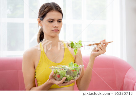 Young woman sitting on sofa eating vegetable salad while feeling dislike and expression disgusted. Young woman sitting on sofa eating vegetable salad while feeling dislike and expression disgusted. 95380713