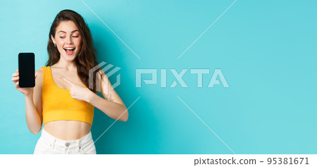 Young happy beautiful girl, pointing and looking at empty smartphone screen, demonstrate advertisement on mobile display, standing against blue background 95381671