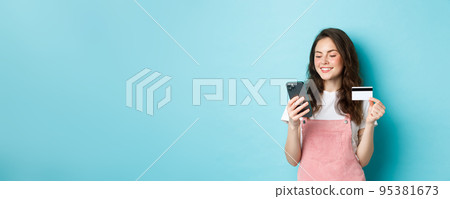 Young modern girl paying online with credit card, type info on smartphone, looking at screen with pleased face, shopping in app, standing over blue background 95381673