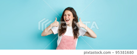 Portrait of smiling cheerful young woman with curly hairstyle, showing thumbs up in approval, say yes, give recommendation, praise good thing, standing against blue background Portrait of smiling cheerful young woman with curly hairstyle, showing thumbs up in approval, say yes, give recommendation, praise good thing, standing against blue background 95381680