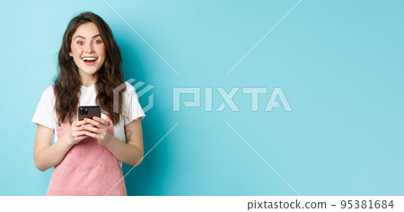 Portrait of excited brunette girl looking amazed at camera while using smartphone, holding mobile phone and smiling cheerful, standing against blue background Portrait of excited brunette girl looking amazed at camera while using smartphone, holding mobile phone and smiling cheerful, standing against blue background 95381684