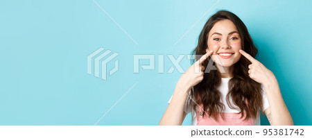 Portrait of happy young woman showing her clean glowing skin and blush on cheeks, pointing fingers at face, smiling satisfied at camera, blue background Portrait of happy young woman showing her clean glowing skin and blush on cheeks, pointing fingers at face, smiling satisfied at camera, blue background 95381742
