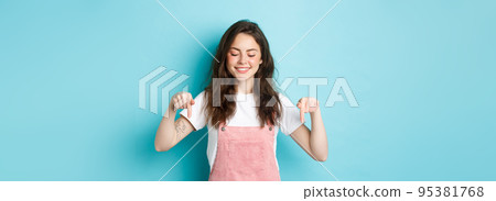 Cute caucasian girl in dungarees pointing and looking down, smiling joyful, showing advertisement on bottom copy space, standing over blue background Cute caucasian girl in dungarees pointing and looking down, smiling joyful, showing advertisement on bottom copy space, standing over blue background 95381768