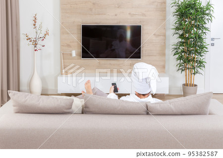 Woman sitting on a couch with towel on her head watching tv Woman sitting on a couch with towel on her head watching tv 95382587