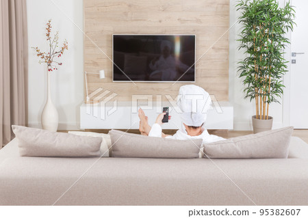 Woman sitting on a couch with towel on her head watching tv Woman sitting on a couch with towel on her head watching tv 95382607