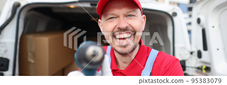 Happy courier in a red uniform shows a tool near a covered van Happy courier in a red uniform shows a tool near a covered van 95383079