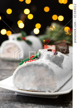 White chocolate yule log cake with christmas lights on wooden table 95384012