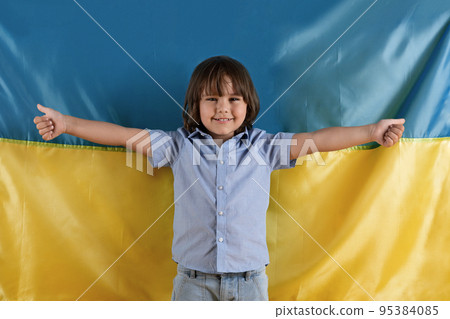 Studio portrait of adorable little boy standing over Ukrainian flag and smiling widely, gesturing thumbs up Studio portrait of adorable little boy standing over Ukrainian flag and smiling widely, gesturing thumbs up 95384085