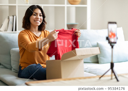 Smiling Arab Female Influencer Unpacking Clothes While Recording Video Content At Home 95384176