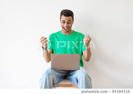 Promotion at work concept. Happy arab man celebrating success looking at laptop, sitting on chair over light wall 95384310