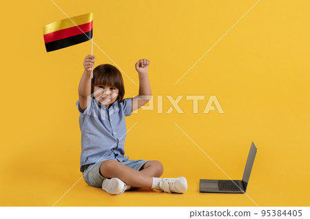 Excited little boy with Germany flag sitting on floor near laptop and smiling, enjoying online education, free space 95384405