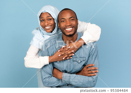Cheerful African American Islamic Spouses Embracing On Blue Studio Background Cheerful African American Islamic Spouses Embracing On Blue Studio Background 95384484