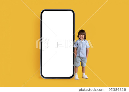 Adorable little boy with hands in pockets standing nera huge smartphone with blank white screen, empty space 95384836