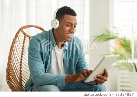 Smiling young african american guy in wireless headphones chatting, searching, surfing in internet Smiling young african american guy in wireless headphones chatting, searching, surfing in internet 95384837