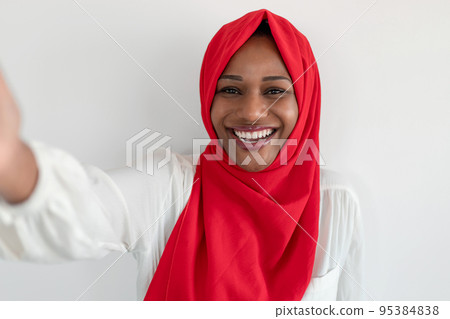 Happy black muslim female blogger making selfie smiling to camera over white wall background, wearing red hijab 95384838