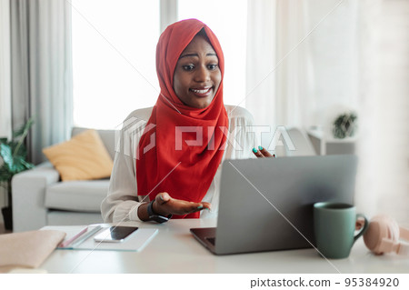African american muslim lady having video call, looking at laptop and gesturing, sitting at workplace in living room 95384920