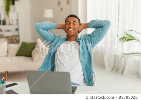 Cheerful millennial african american man with closed eyes resting at work with laptop in living room interior 95385020