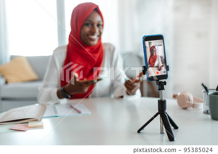 Positive black islamic lady capturing content for vlog at home, recording video on smartphone on tripod, selective focus Positive black islamic lady capturing content for vlog at home, recording video on smartphone on tripod, selective focus 95385024