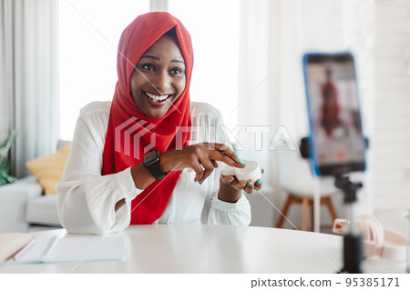Professional black muslim beauty blogger advertising skincare cosmetics, talking to smartphone on tripod 95385171