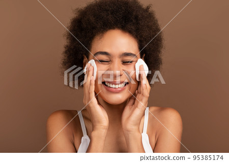 Happy young mixed race lady with curly hair removes makeup from face with cotton pad Happy young mixed race lady with curly hair removes makeup from face with cotton pad 95385174