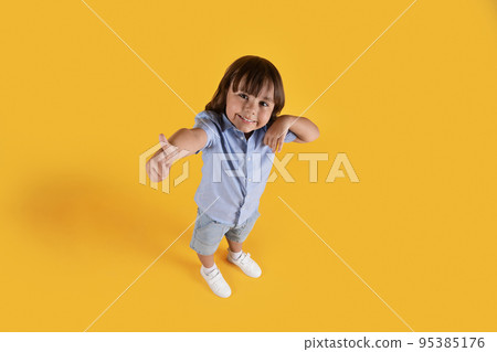 Above view portrait of little boy showing thumb up gesture, smiling to camera, orange studio background, full length Above view portrait of little boy showing thumb up gesture, smiling to camera, orange studio background, full length 95385176