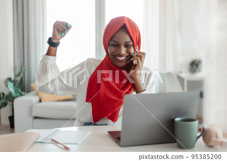 Emotional muslim female entrepreneur talking on cellphone, looking at laptop and gesturing, successful business deal 95385200