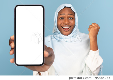 Excited black muslim woman gesturing YES and showing smartphone with white empty screen, mockup Excited black muslim woman gesturing YES and showing smartphone with white empty screen, mockup 95385253