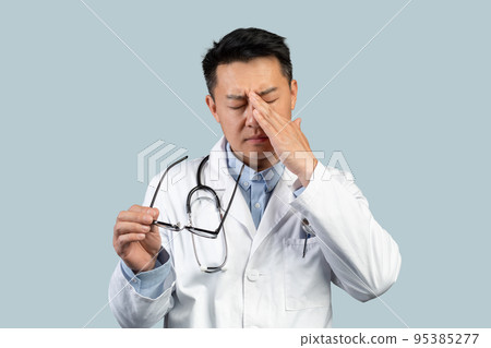 Tired sad middle aged chinese man doctor in white coat takes off glasses, rubs eyes with hand 95385277