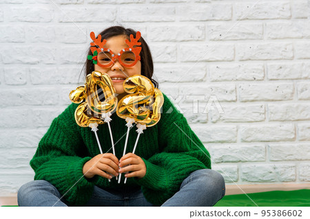 Cheerful Asian child girl in green sweater, masquerade Christmas glasses with balls with number 2023 Cheerful Asian child girl in green sweater, masquerade Christmas glasses with balls with number 2023 95386602