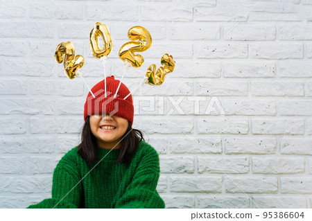 Smiling Asian baby girl in a green sweater close-up with golden balloons with numbers 2023 in hat Smiling Asian baby girl in a green sweater close-up with golden balloons with numbers 2023 in hat 95386604