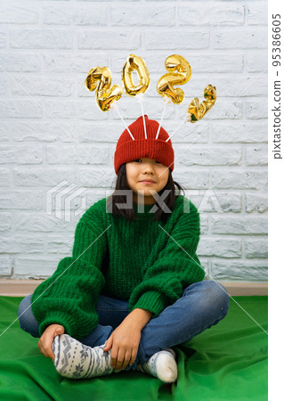 Funny Asian girl in green sweater, sits on floor with golden balloons with number 2023 in a red hat Funny Asian girl in green sweater, sits on floor with golden balloons with number 2023 in a red hat 95386605