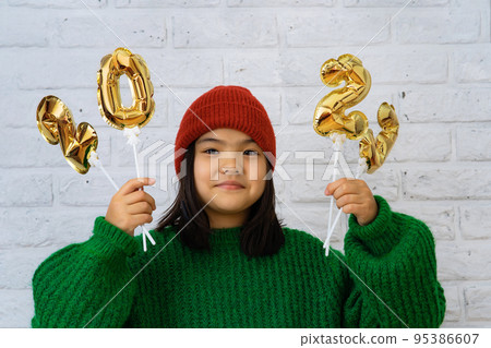 Asian child girl in ahat and green sweater close-up, in hands of golden balloons with numbers 2023 Asian child girl in ahat and green sweater close-up, in hands of golden balloons with numbers 2023 95386607