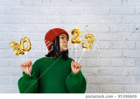 Asian cute girl in red cap and green sweater holding foil balloons with numbers 2023. Asian cute girl in red cap and green sweater holding foil balloons with numbers 2023. 95386609