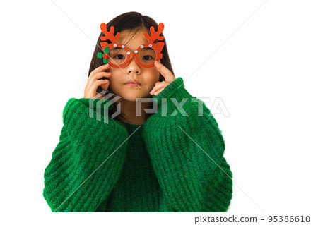 Beautiful Asian child girl in a green sweater puts on masquerade Christmas glasses with deer horns Beautiful Asian child girl in a green sweater puts on masquerade Christmas glasses with deer horns 95386610
