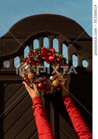 Women's hands decorate entrance wooden door with festive Christmas wreath Women's hands decorate entrance wooden door with festive Christmas wreath 95386614