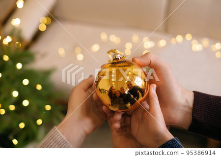 A Christmas golden ball in the hands with a reflection of the family. Cheerful parents with their sons are holding a toy. Time to decorate the Christmas tree A Christmas golden ball in the hands with a reflection of the family. Cheerful parents with their sons are holding a toy. Time to decorate the Christmas tree 95386914