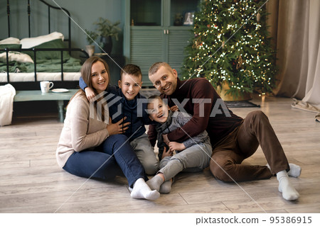 The family is having fun and cuddling together sitting on the floor in a room with a Christmas tree. A house decorated with Christmas decor and lights The family is having fun and cuddling together sitting on the floor in a room with a Christmas tree. A house decorated with Christmas decor and lights 95386915