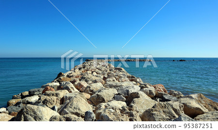 Breakwater of large stones in blue sea to horizon, road to sea, beautiful seascape, Mediterranean beaches, holidays in Spain 95387251