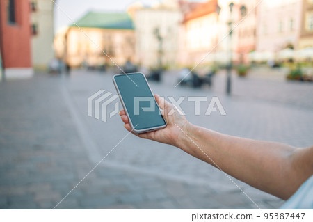 Close up of women's hands holding cell telephone with blank copy space scree for your advertising text message or promotional content, hipster girl watching video on mobile phone in Krakow, Poland Close up of women's hands holding cell telephone with blank copy space scree for your advertising text message or promotional content, hipster girl watching video on mobile phone in Krakow, Poland 95387447