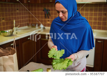 Middle-Eastern Muslim woman, a pretty housewife with head covered in blue hijab, holds fresh salad leaves in her hands while unpacking grocery bag in the home kitchen, after shopping. Food delivery. Middle-Eastern Muslim woman, a pretty housewife with head covered in blue hijab, holds fresh salad leaves in her hands while unpacking grocery bag in the home kitchen, after shopping. Food delivery. 95387707