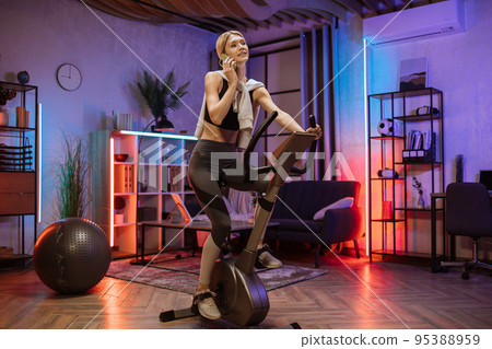 Beautiful sporty fit female cycling stationary bike and watching online fitness class on smartphone. Beautiful sporty fit female cycling stationary bike and watching online fitness class on smartphone. 95388959
