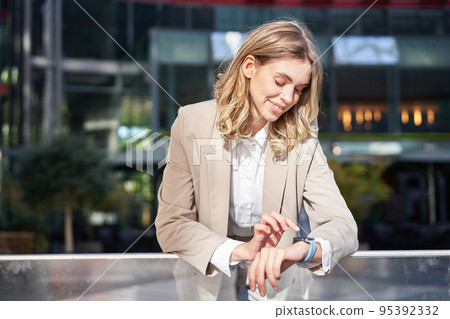 Smiling businesswoman looks at her digital watch, reads message or checks time, stands outside on street in beige office suit 95392332