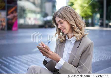 Young businesswoman preparing for speech, interview in company, writing down notes while sitting outside in city centre 95392333