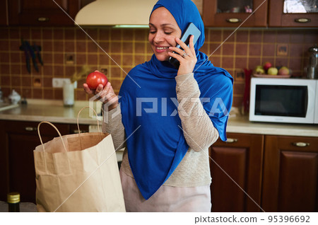 Charming Muslim Middle-Eastern woman in hijab, housewife talking on the phone while arriving home after grocery shopping, unpacking bags, taking out fresh vegetables and groceries. Food delivery Charming Muslim Middle-Eastern woman in hijab, housewife talking on the phone while arriving home after grocery shopping, unpacking bags, taking out fresh vegetables and groceries. Food delivery 95396692