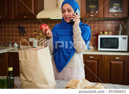 Pleasant housewife, Arab Muslim woman in blue hijab, returning home from grocery store, talking on mobile phone, taking fresh vegetables out of bags, unpacking groceries in the kitchen. Food delivery Pleasant housewife, Arab Muslim woman in blue hijab, returning home from grocery store, talking on mobile phone, taking fresh vegetables out of bags, unpacking groceries in the kitchen. Food delivery 95396693